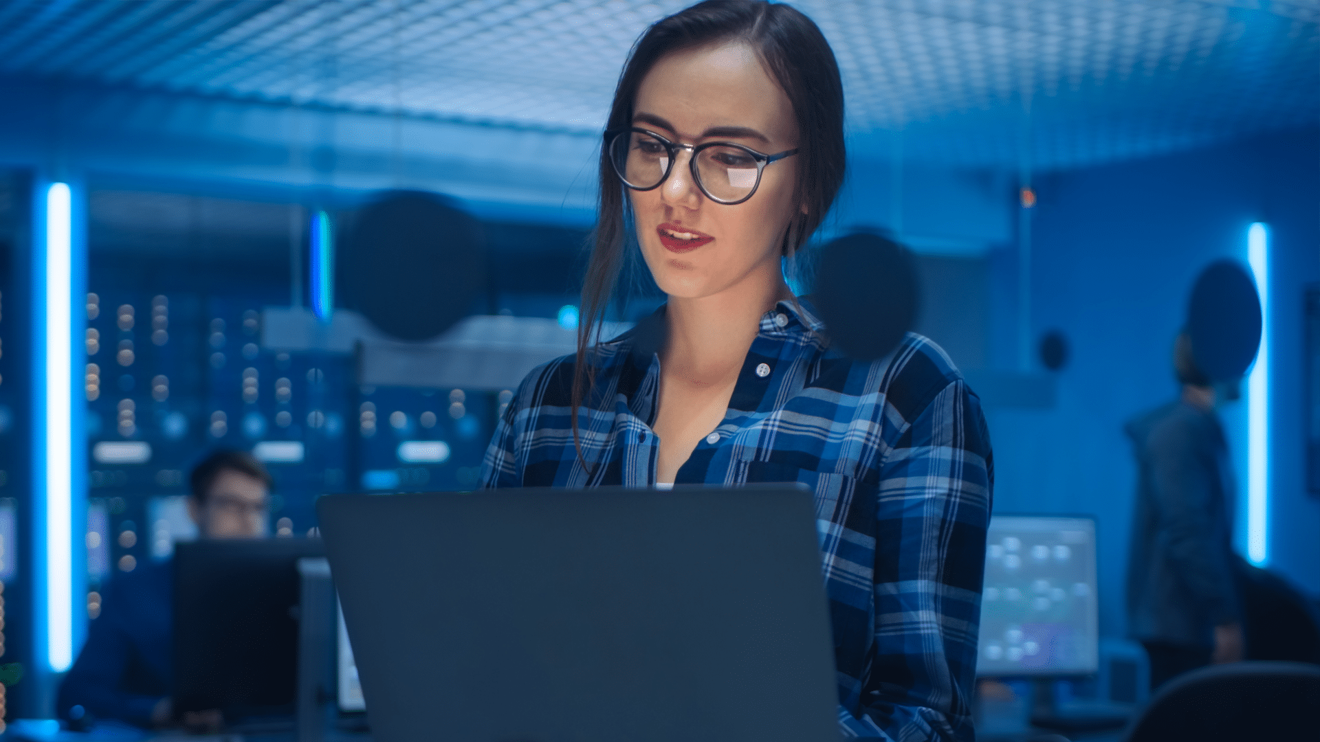 woman wearing glasses working on a laptop with male colleagues in a data center.